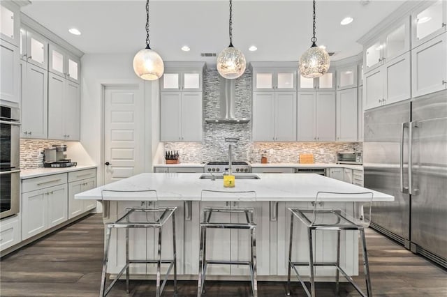 kitchen featuring built in fridge, light stone countertops, dark hardwood / wood-style flooring, an island with sink, and wall chimney range hood