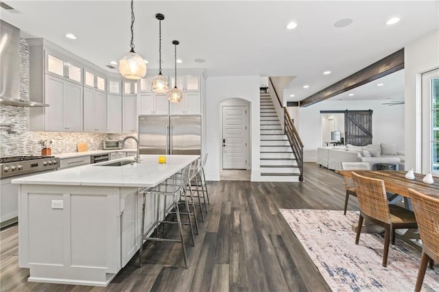 kitchen featuring stainless steel built in fridge, an island with sink, dark wood-type flooring, sink, and wall chimney range hood