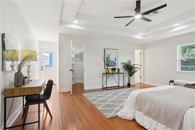 bedroom with ceiling fan, a walk in closet, light wood-type flooring, and a tray ceiling