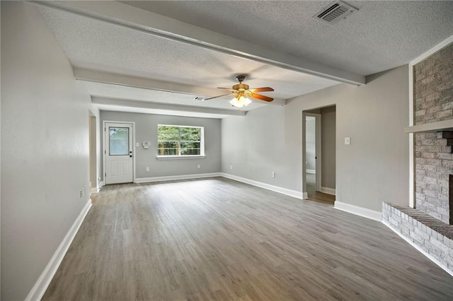 unfurnished living room with a textured ceiling, beamed ceiling, wood-type flooring, a brick fireplace, and ceiling fan