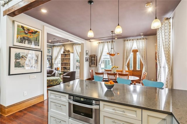 kitchen with dark stone counters, hanging light fixtures, stainless steel oven, dark wood-type flooring, and cream cabinetry