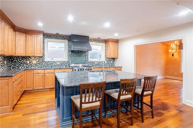 kitchen featuring an island with sink, light wood-type flooring, stone countertops, and wall chimney range hood