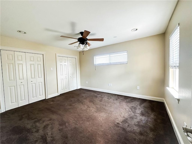 unfurnished bedroom featuring multiple windows, dark carpet, ceiling fan, and multiple closets