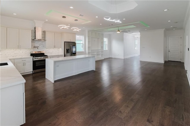 kitchen featuring ceiling fan with notable chandelier, dark hardwood / wood-style flooring, stainless steel appliances, a center island, and wall chimney range hood