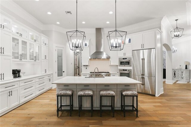 kitchen featuring stainless steel refrigerator, wall chimney exhaust hood, a kitchen island with sink, and hanging light fixtures