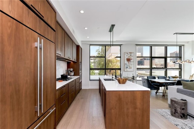 kitchen with sink, light wood-type flooring, an island with sink, hanging light fixtures, and paneled fridge