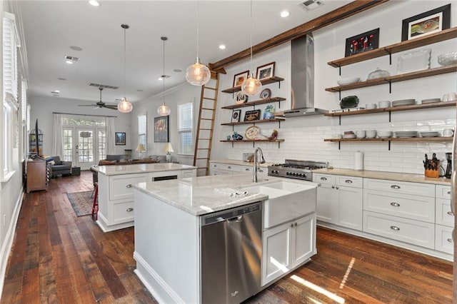 kitchen featuring an island with sink, white cabinetry, wall chimney range hood, stainless steel appliances, and decorative light fixtures