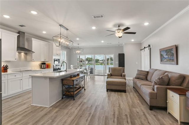 kitchen with white cabinetry, a barn door, light wood-type flooring, a kitchen island with sink, and wall chimney range hood