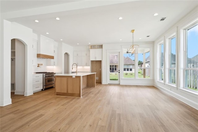 kitchen with pendant lighting, light wood-type flooring, a kitchen island with sink, stainless steel stove, and white cabinetry