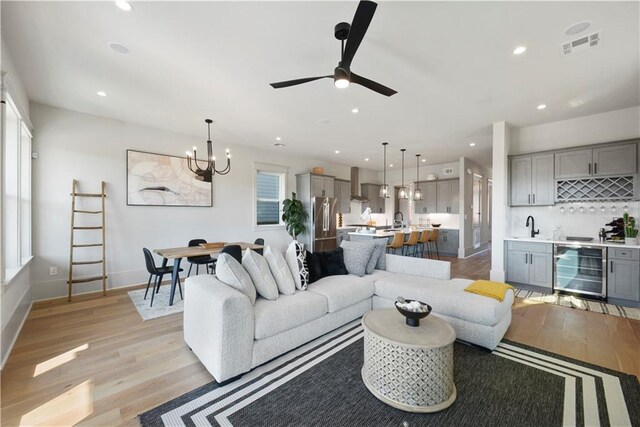 living room featuring beverage cooler, ceiling fan with notable chandelier, sink, and light hardwood / wood-style flooring