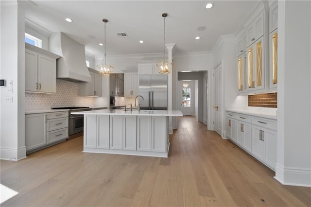 kitchen featuring appliances with stainless steel finishes, light hardwood / wood-style floors, pendant lighting, custom exhaust hood, and a kitchen island with sink