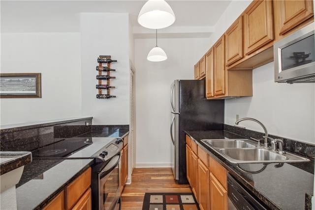 kitchen with appliances with stainless steel finishes, hanging light fixtures, sink, dark stone countertops, and light hardwood / wood-style flooring