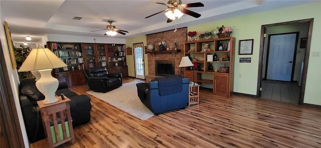 living room with hardwood / wood-style floors, a brick fireplace, a tray ceiling, and ceiling fan