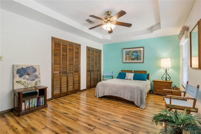 bedroom with light hardwood / wood-style floors, multiple closets, a raised ceiling, and ceiling fan