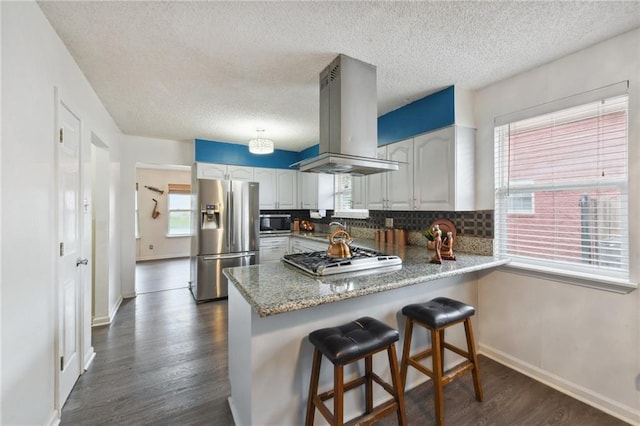 kitchen with kitchen peninsula, tasteful backsplash, wall chimney exhaust hood, white cabinetry, and stainless steel appliances