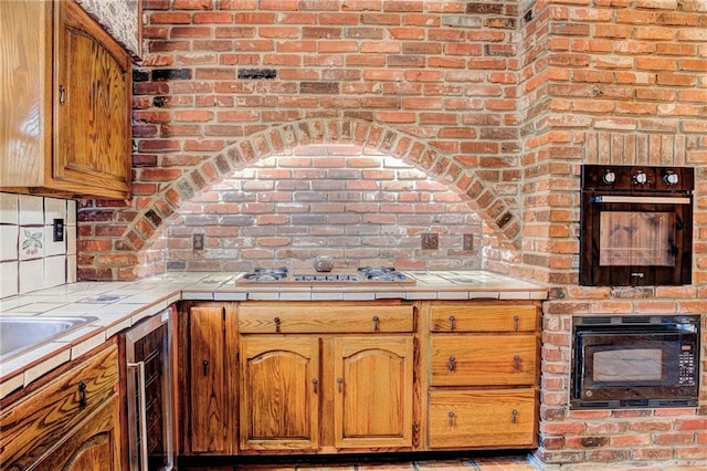 kitchen featuring tile counters, black microwave, beverage cooler, stainless steel gas stovetop, and brick wall