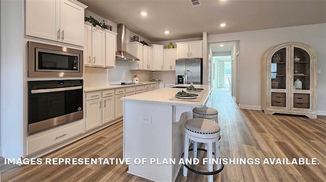 kitchen with white cabinets, wall chimney range hood, a breakfast bar area, light hardwood / wood-style flooring, and stainless steel appliances