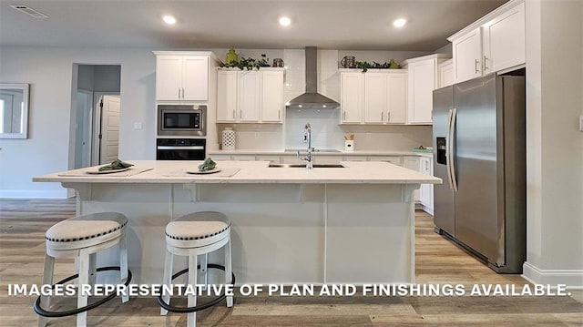 kitchen with a center island with sink, wall chimney range hood, light hardwood / wood-style flooring, appliances with stainless steel finishes, and white cabinetry