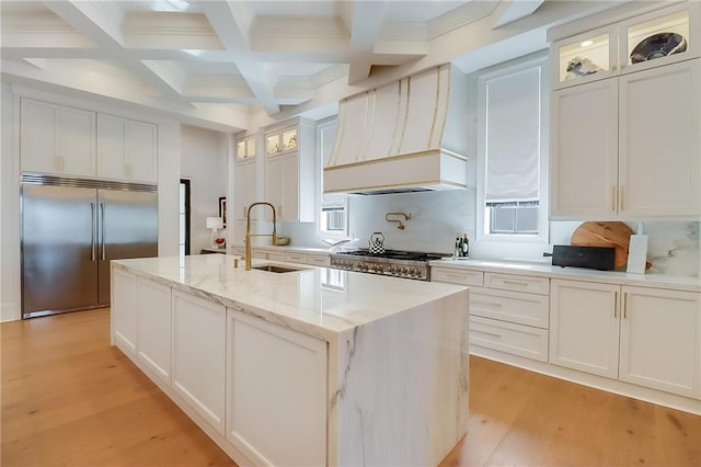 kitchen with a kitchen island with sink, custom range hood, sink, white cabinetry, and appliances with stainless steel finishes