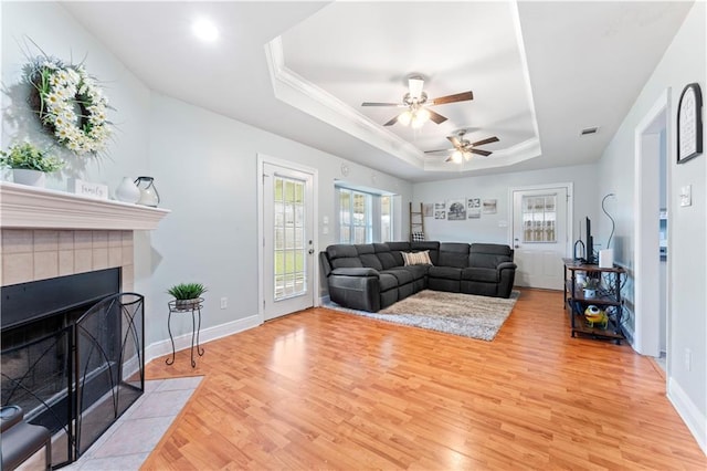 living room featuring ceiling fan, a tray ceiling, a tiled fireplace, and light hardwood / wood-style floors