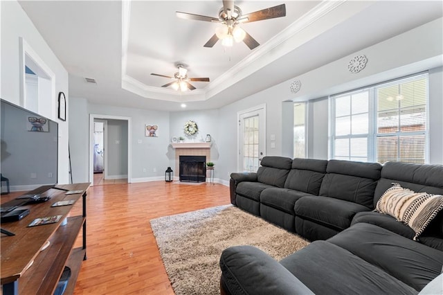 living room featuring light hardwood / wood-style floors, ceiling fan, a tiled fireplace, a tray ceiling, and crown molding