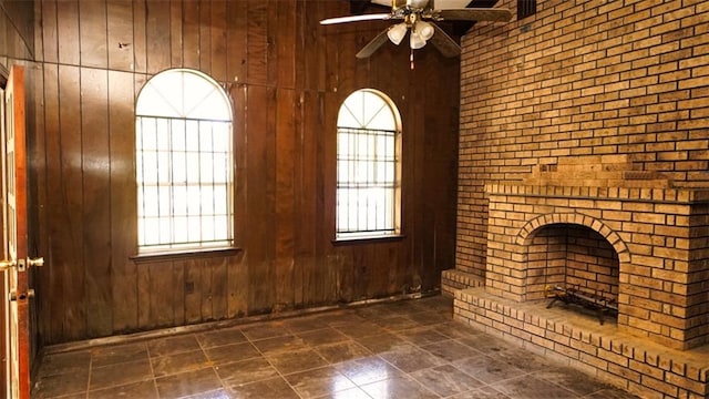 unfurnished living room featuring ceiling fan, a brick fireplace, wood walls, and a wealth of natural light