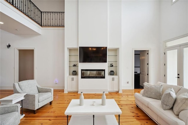 living area featuring light wood-style flooring, a glass covered fireplace, french doors, a high ceiling, and baseboards