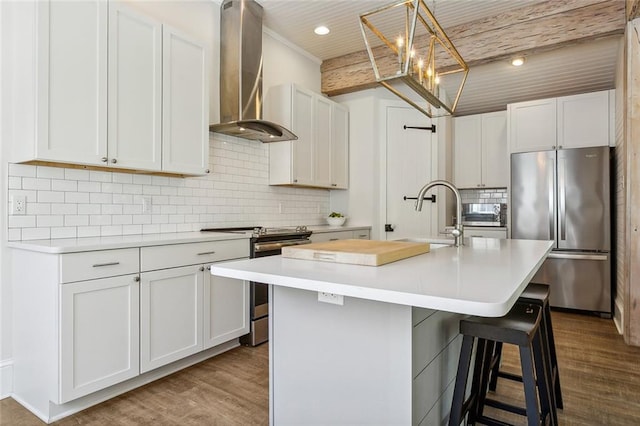 kitchen with wall chimney range hood, a kitchen island with sink, white cabinets, appliances with stainless steel finishes, and hardwood / wood-style flooring