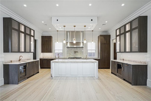 kitchen with light wood-type flooring, wall chimney range hood, hanging light fixtures, a spacious island, and wine cooler