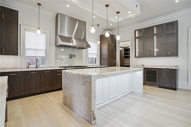 kitchen with dark brown cabinetry, a raised ceiling, wall chimney exhaust hood, ornamental molding, and a sink