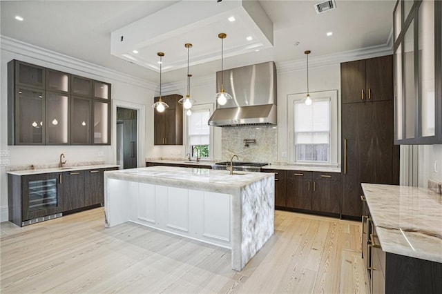 kitchen featuring a tray ceiling, light wood-style floors, wall chimney range hood, dark brown cabinets, and beverage cooler