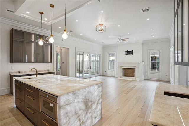 kitchen with light hardwood / wood-style floors, pendant lighting, dark brown cabinets, a center island with sink, and light stone countertops