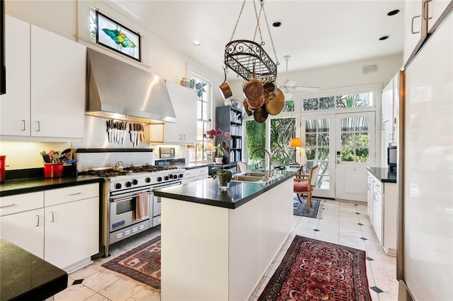 kitchen featuring double oven range, white cabinets, sink, wall chimney exhaust hood, and an island with sink