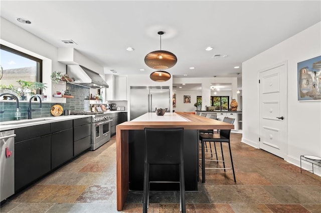 kitchen featuring high end appliances, sink, pendant lighting, wall chimney range hood, and backsplash