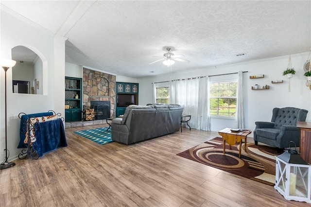 living room with ceiling fan, a stone fireplace, hardwood / wood-style flooring, crown molding, and a textured ceiling