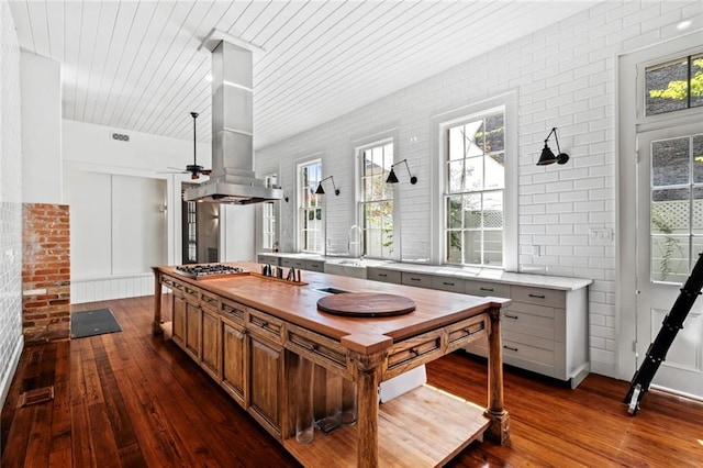 kitchen with island exhaust hood, ceiling fan, dark hardwood / wood-style flooring, stainless steel gas stovetop, and brick wall