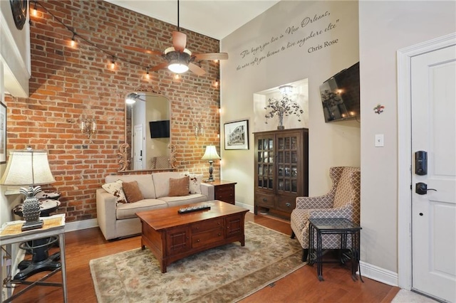 living area featuring wood finished floors, brick wall, baseboards, ceiling fan, and a high ceiling