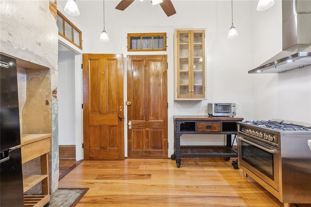 kitchen featuring wall chimney range hood, light wood-type flooring, ceiling fan, decorative light fixtures, and high end stove