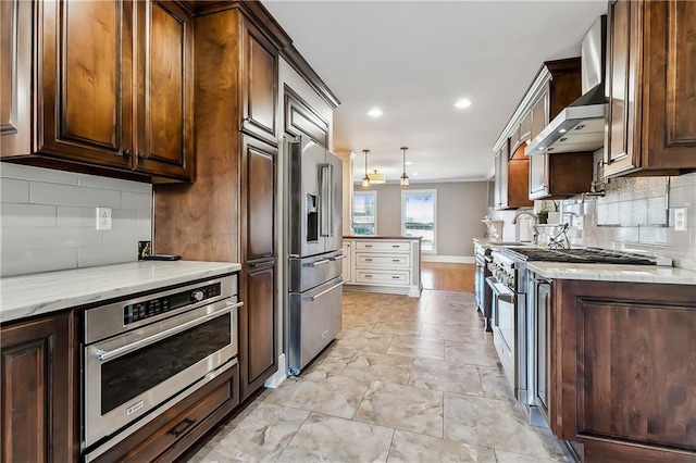 kitchen featuring premium appliances, wall chimney range hood, sink, tasteful backsplash, and pendant lighting