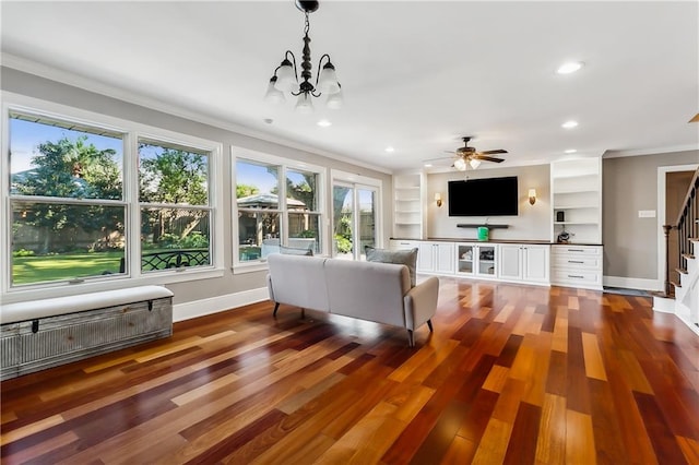 living room with ornamental molding, dark wood-type flooring, built in shelves, and ceiling fan with notable chandelier