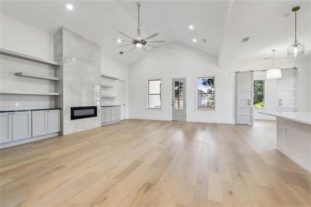 unfurnished living room featuring ceiling fan, a barn door, high vaulted ceiling, light wood-type flooring, and a fireplace