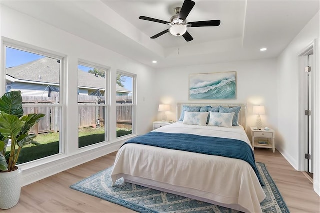 bedroom featuring ceiling fan, a tray ceiling, multiple windows, and light wood-type flooring