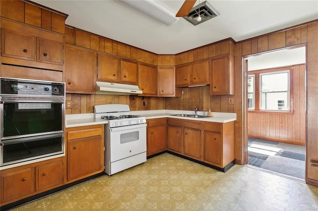 kitchen with ceiling fan, sink, double oven, wood walls, and white gas range oven