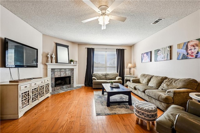 living room with light hardwood / wood-style flooring, a tiled fireplace, a textured ceiling, and ceiling fan