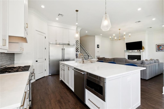 kitchen with dark hardwood / wood-style floors, built in appliances, an island with sink, white cabinets, and decorative light fixtures