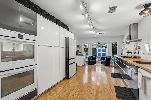 kitchen with white cabinets, ceiling fan, appliances with stainless steel finishes, light wood-type flooring, and wall chimney exhaust hood