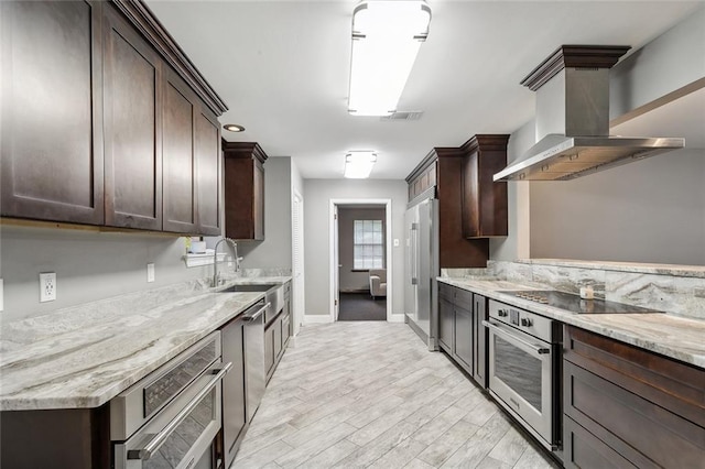 kitchen featuring wall chimney range hood, dark brown cabinets, stainless steel appliances, light stone counters, and light hardwood / wood-style flooring