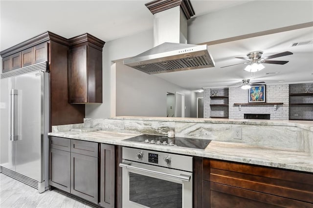 kitchen with range hood, dark brown cabinetry, appliances with stainless steel finishes, light stone counters, and a fireplace