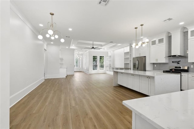 kitchen with appliances with stainless steel finishes, ceiling fan with notable chandelier, a tray ceiling, wall chimney range hood, and white cabinets