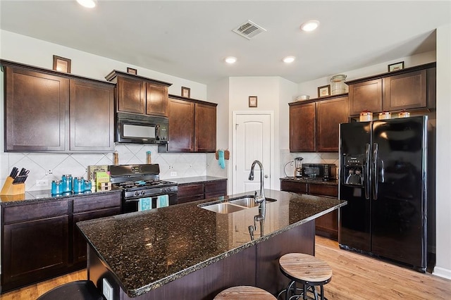 kitchen featuring light hardwood / wood-style flooring, dark stone countertops, a center island with sink, sink, and black appliances